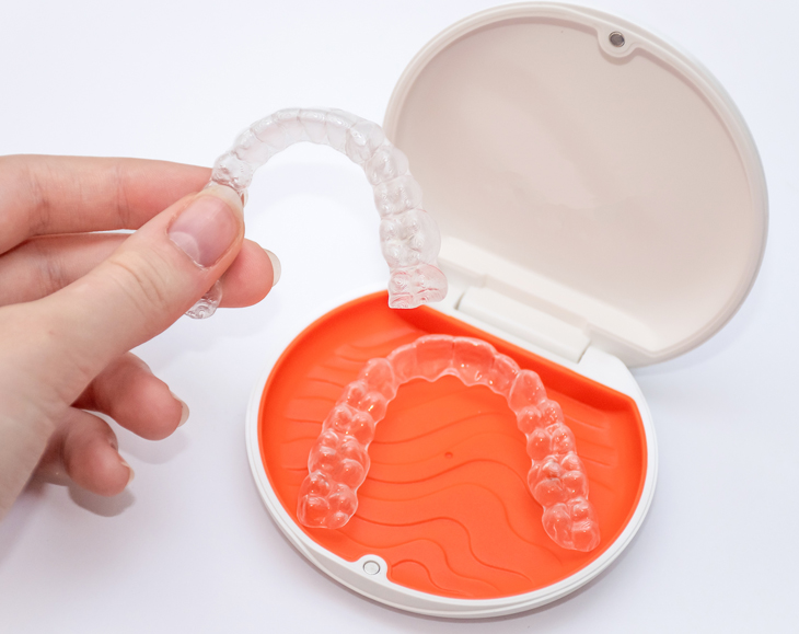 Hand holding open clear case displaying orange orthodontic retainer model against white background.