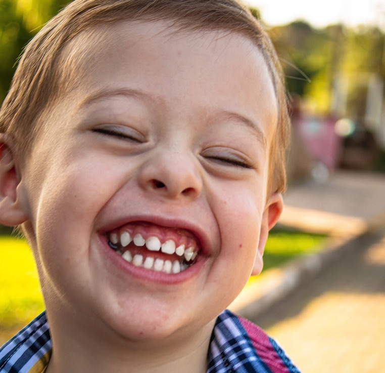 Young child smiling brightly outdoors in checkered shirt, displaying healthy teeth.