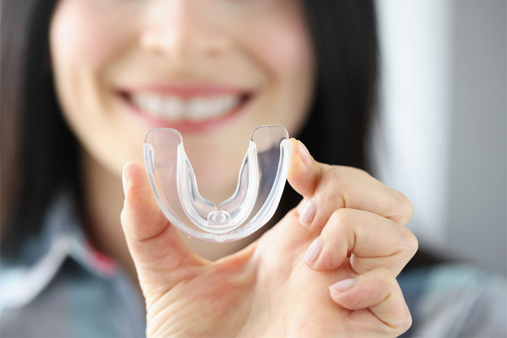 Smiling woman holding clear orthodontic retainer or aligner in front of blurred background.