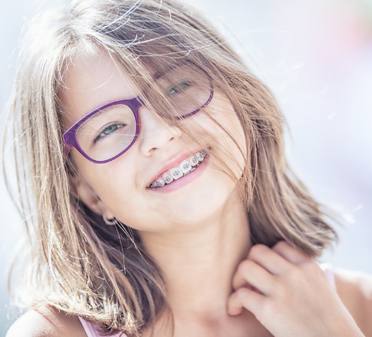 Young girl wearing purple glasses smiling brightly at camera outdoors.