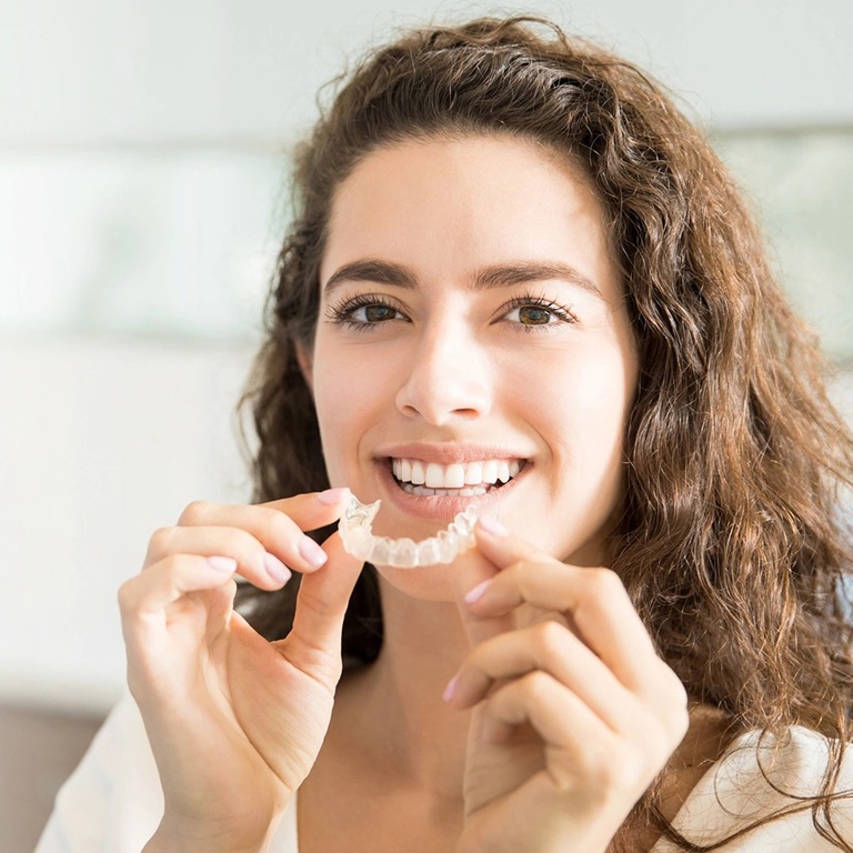 Woman holding clear aligner trays, smiling indoors in dental office setting.