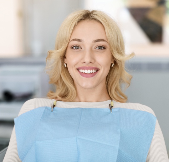 Professional female dentist smiling at camera in modern dental office setting wearing blue scrubs.